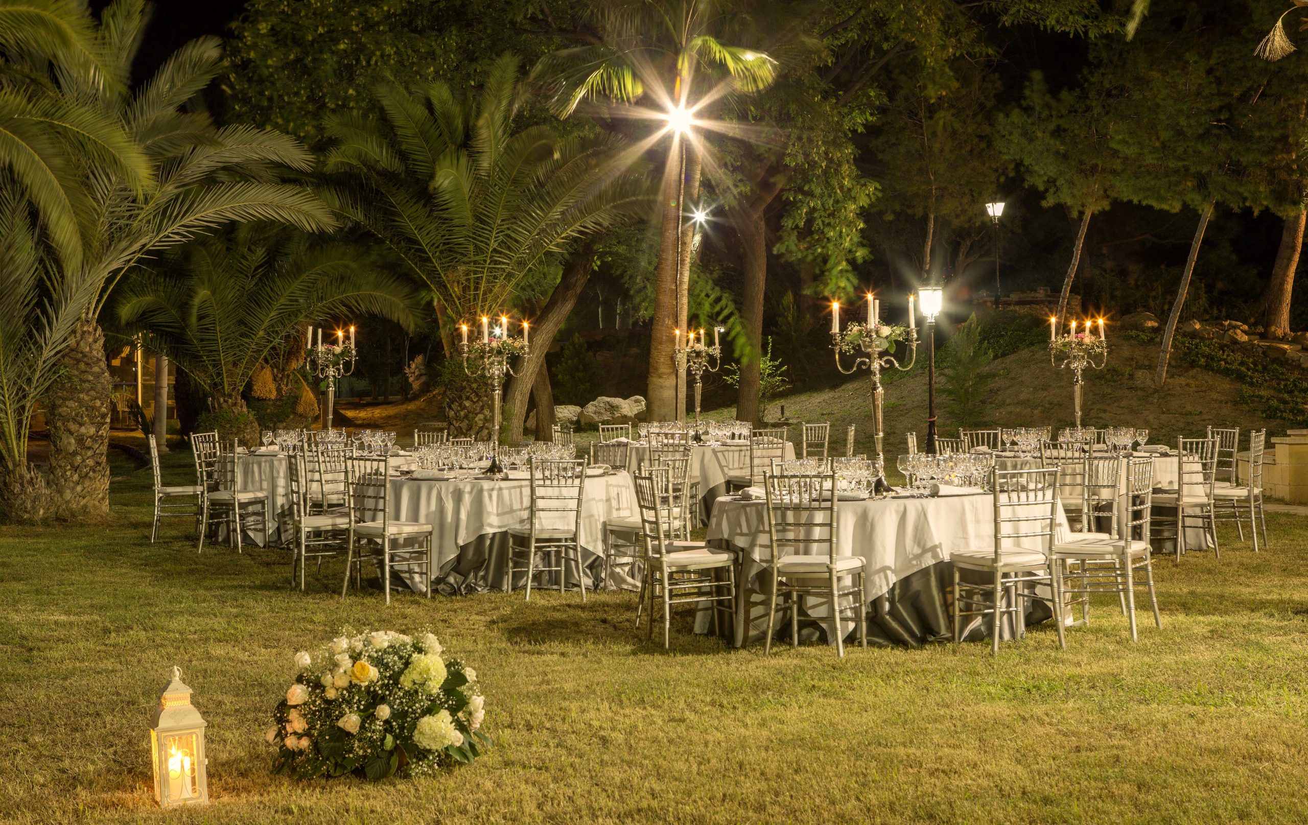 Salón exterior iluminado con candelabros altos y farolas elegantes entre palmeras.